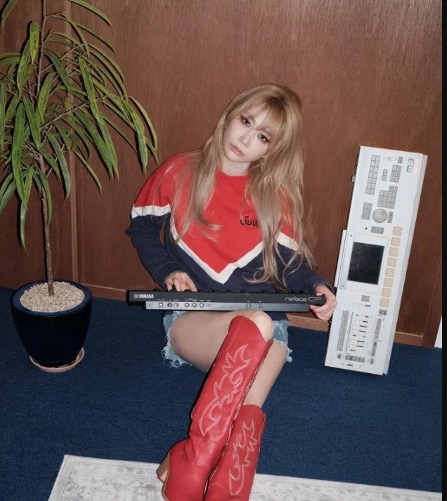 JeA sits on a blue carpet in red cowboy boots, holding a Yamaha keyboard beside music gear.