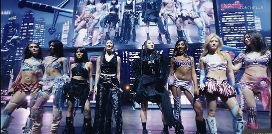 KATSEYE performs on stage at Coachella with members in bold outfits, dancing under large screens and stage lights.