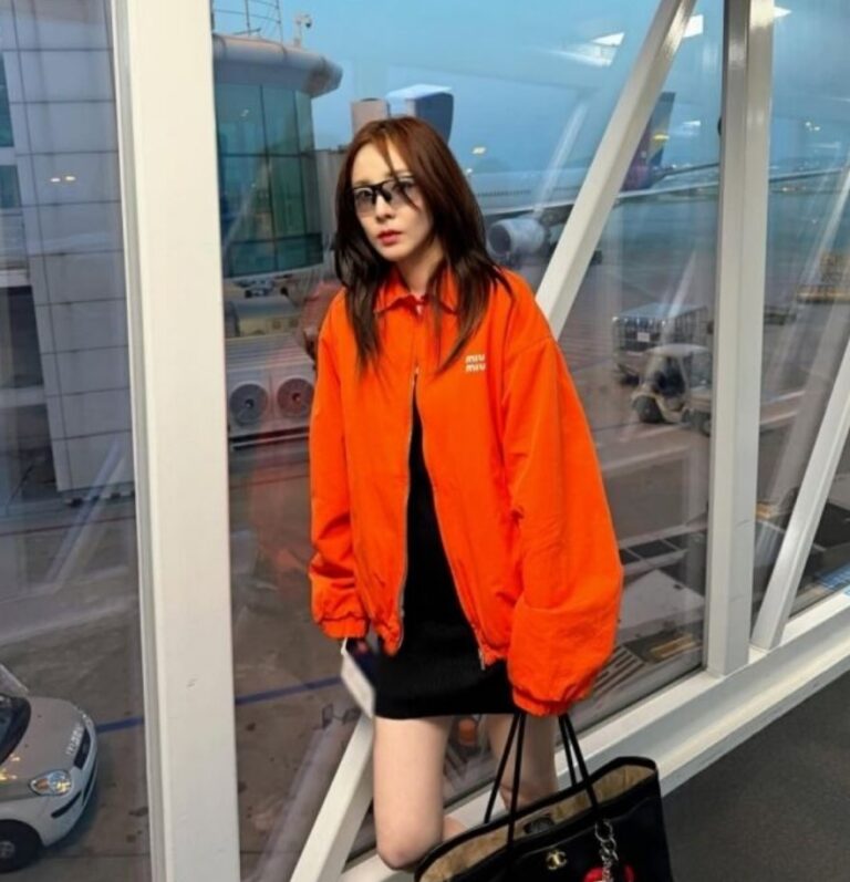 Sandara Park walks through an airport jet bridge and terminal, wearing a bright orange MIU MIU jacket and carrying a Chanel bag, showcasing her chic travel fashion.
