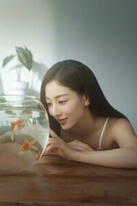 Jihyo leans on a table, gazing at goldfish swimming in a round glass bowl.