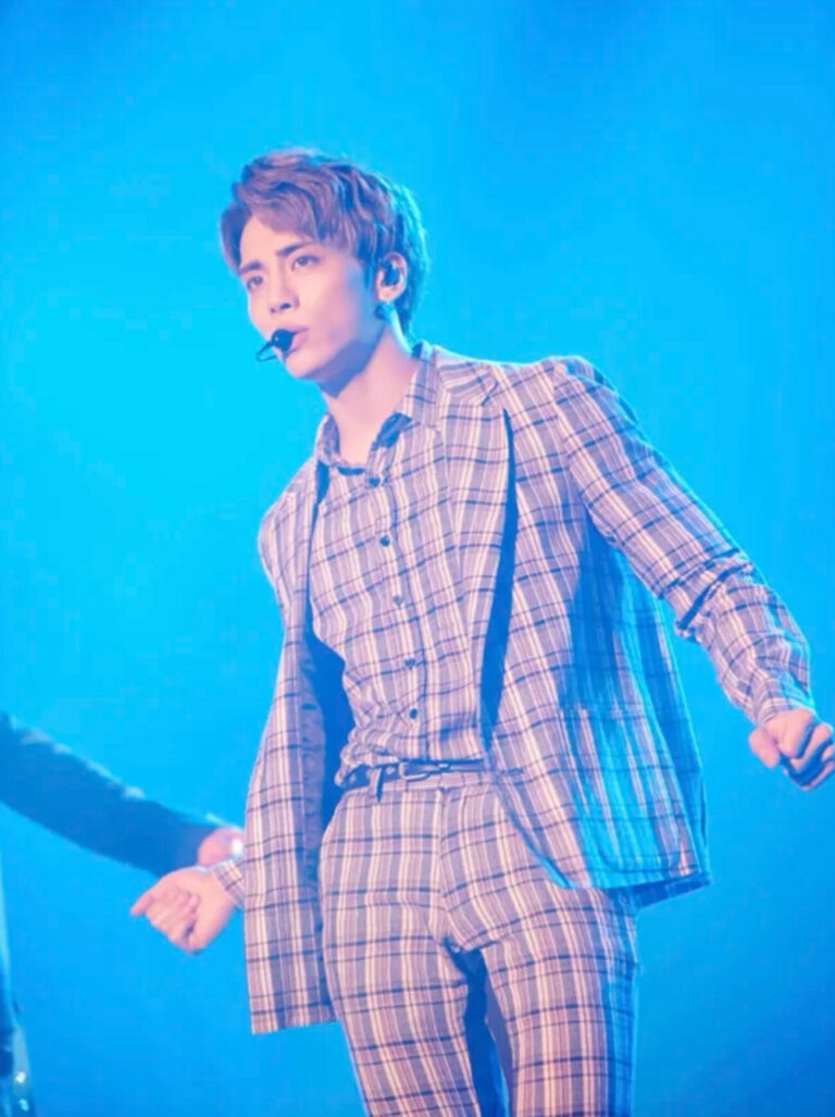 Kim Jong-hyun performing on stage in a plaid suit with a headset mic under blue lighting.