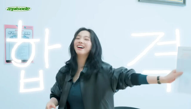 Karina holds a paper with glowing “합격” text, celebrating success indoors.