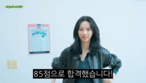 Karina stands against a wall with a poster behind her, smiling with text “85점으로 합격했습니다!”