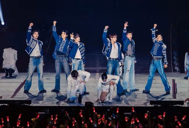 2PM perform energetically on stage in blue and white outfits, raising their arms as red light sticks glow across the crowd.