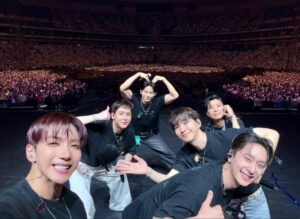 2PM members smile and pose on stage at Tokyo Dome, surrounded by a massive cheering crowd with glowing lights.