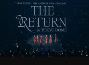 2PM members stand on stage at Tokyo Dome in matching red coats under a starry backdrop reading “THE RETURN,” celebrating their 15th Japan debut anniversary concert.