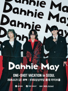 Three members of Dannie May stand against a backdrop repeating their band name, dressed in dark, stylish suits with bold red accents.