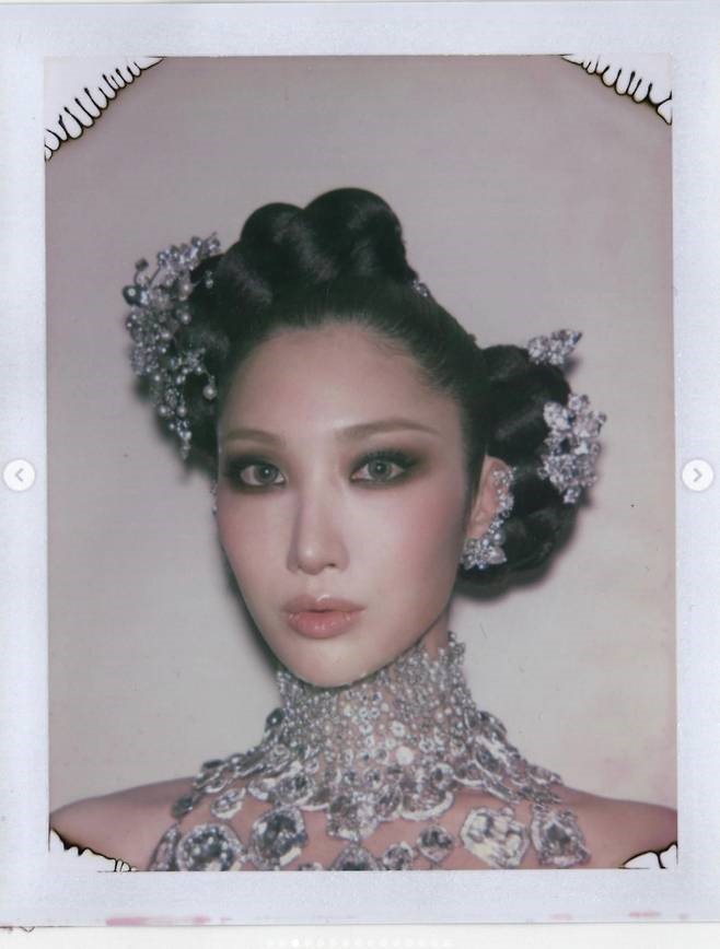 Close‑up portrait of EJ wearing an ornate jeweled collar and crystal hair accessories, styled in sculpted waves with dramatic smoky eye makeup.