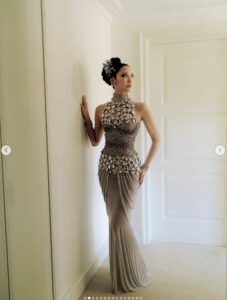EJ poses in a full‑length, beaded gown with cascading crystal chains and matching hair ornaments, standing against a cream‑colored wall.