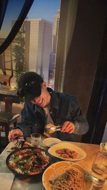 Bang Chan smiles while dining at a restaurant table with pasta and salad dishes.