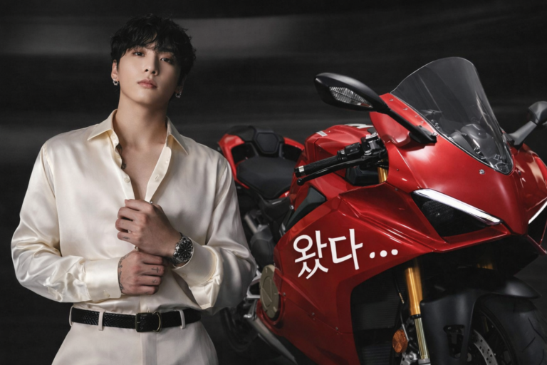 BTS’s Jungkook poses confidently beside his new red Ducati motorcycle, wearing a cream silk shirt and watch, with Korean text “왔다…” visible on the bike.