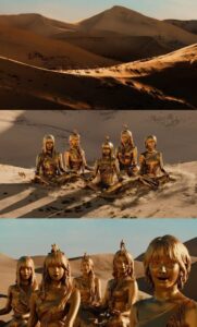 LE SSERAFIM appears as golden statues in a vast desert landscape, surrounded by sand dunes and warm sunlight, blending surreal and cinematic imagery.