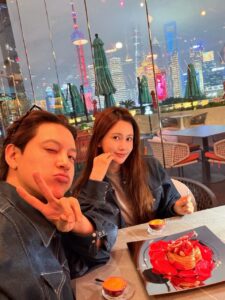 Lee Da‑hae and SE7EN pose at a restaurant with a view of Shanghai’s illuminated skyline, smiling over dessert.