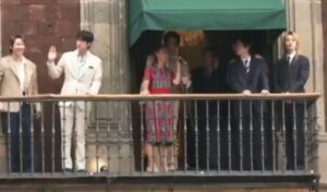 BTS members stand on a balcony at Mexico’s presidential palace, waving and greeting the public.