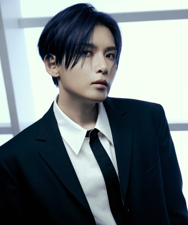 Ryeowook in a formal black suit with neat hairstyle, posing against a softly lit background.