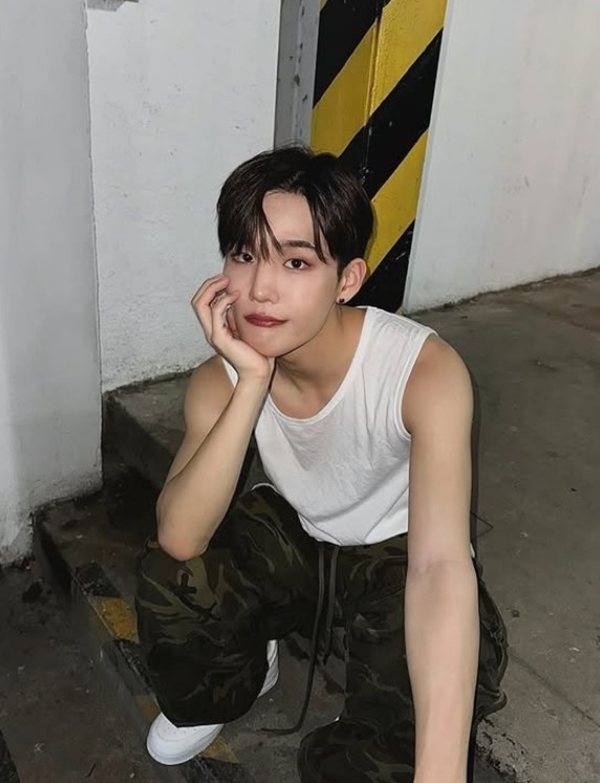 Seunghun sits in a dimly lit urban setting wearing a sleeveless shirt and camouflage pants.