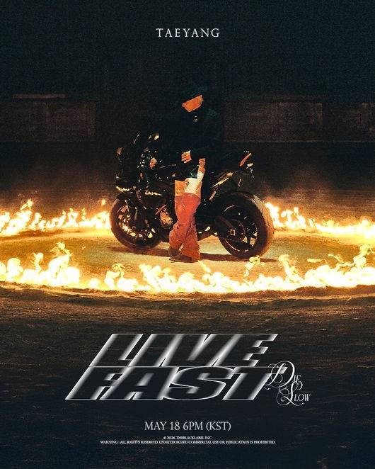 Taeyang stands beside a black motorcycle encircled by flames in a dark setting, with bold metallic text reading “LIVE FAST DIE SLOW.”