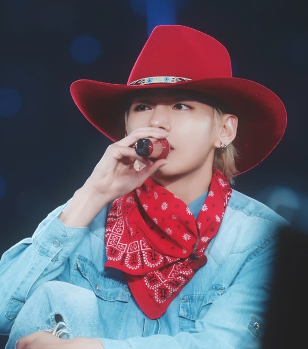 BTS member V performs wearing a red cowboy hat, matching bandana, and denim outfit, holding a microphone during a concert with blue lights in the background.