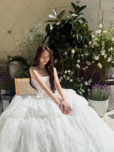 Yoona sits outdoors surrounded by lush flowers, wearing a voluminous white lace gown and silver jewelry, smiling serenely in natural sunlight.