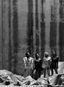 aespa members stand in avant-garde outfits before a tall, textured concrete wall, surrounded by draped metallic fabric in a monochrome setting.