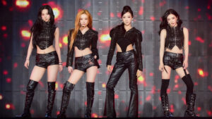 aespa members performing in edgy black leather outfits under red stage lights.