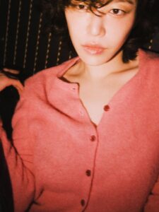 Ga In wearing a pink cardigan against a dark background with warm lighting.