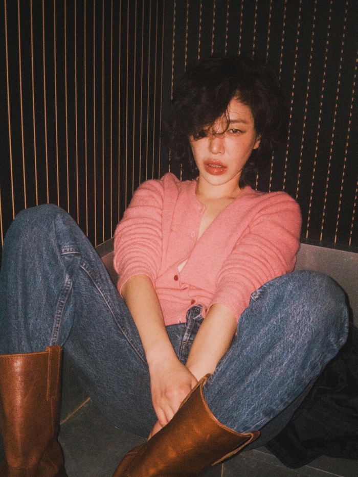 Ga In sitting casually on the floor in a pink cardigan, jeans, and boots, in a dimly lit corner.