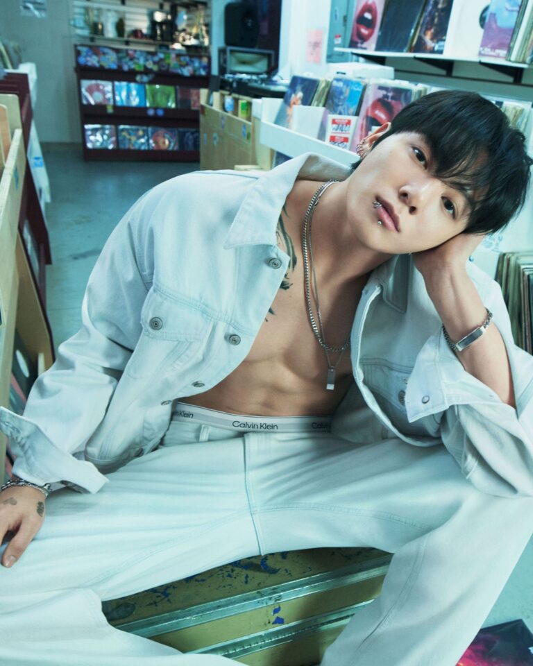Jungkook sits in a record store wearing light denim, revealing Calvin Klein underwear and silver jewelry, surrounded by vinyl albums.