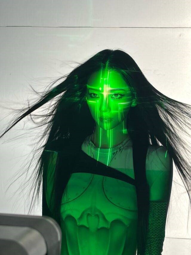 Karina from aespa stands against a light background with long hair flowing, illuminated by bright green laser projections forming futuristic patterns across her face and body.