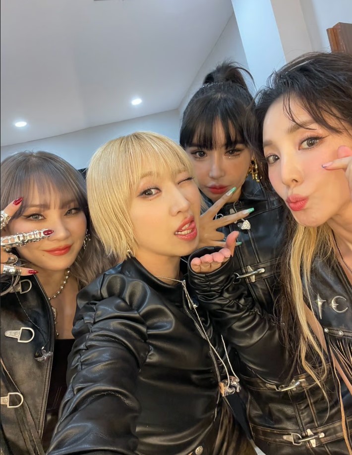 2NE1 members Minzy, Bom, CL, and Dara pose together in black leather outfits, smiling and making playful gestures in a brightly lit room.