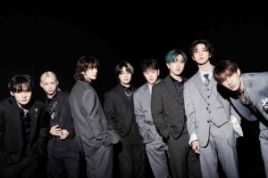 Stray Kids members stand in a row wearing tailored dark suits against a black background, exuding a polished and confident aura.