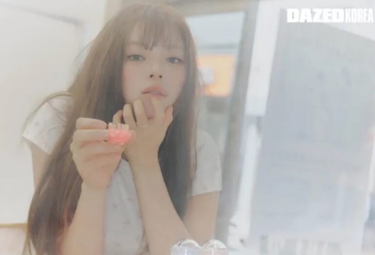 Jeongyeon sits indoors with long brown hair, holding a small pink cosmetic item and gazing softly at the camera in a hazy, pastel-toned setting.