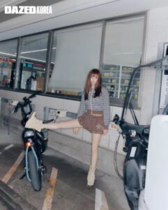 Jeongyeon stands in a gas station setting, wearing a striped top, brown shorts, and beige boots, playfully resting one leg on a motorcycle.