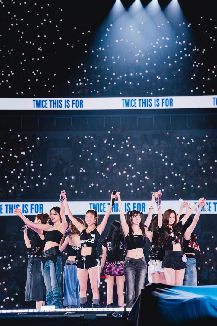 TWICE’s nine members stand on stage in coordinated black and denim outfits, celebrating with fans.