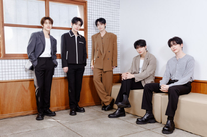 ZEROBASEONE members pose indoors in neutral-toned outfits, seated and standing against a tiled wall and wooden window, exuding calm confidence.