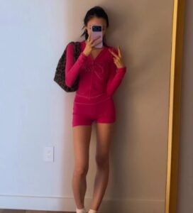 Giselle takes a mirror selfie indoors wearing a bright pink zip‑up top and matching shorts, paired with a leopard‑print bag.