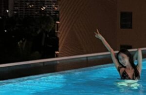Giselle poses joyfully in a rooftop pool at night, raising one arm as city lights shimmer in the background.