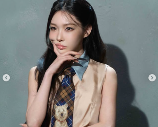 Chungha poses against a gray wall wearing a sleeveless beige top, blue collared shirt, and plaid tie featuring a small dog design.