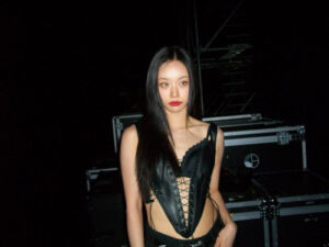 Hyeri stands backstage in a dimly lit area, wearing a black lace-up corset top and black pants. Her straight hair and bold red lipstick create a striking look against the dark background and equipment cases.