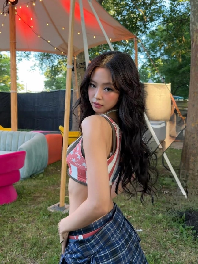 BLACKPINK’s Jennie poses outdoors at Governors Ball in New York, wearing a red, white, and blue crop top with a plaid shirt tied around her waist. The background shows colorful lounge chairs and a tent decorated with string lights.