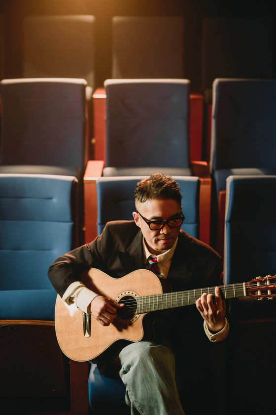 Singer Kim Gun-mo sits among theater seats, playing an acoustic guitar under warm lighting, focused and calm.