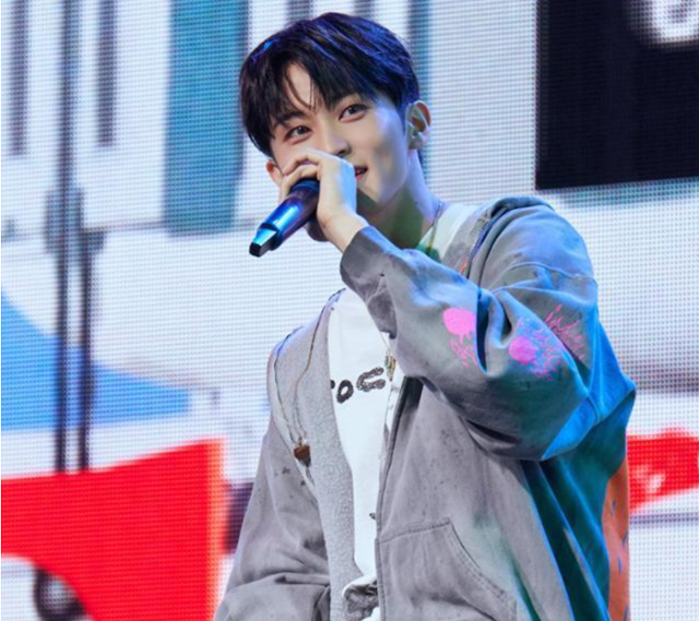 Former NCT member Mark performs on stage wearing a gray hoodie and holding a microphone, smiling under blue stage lights.