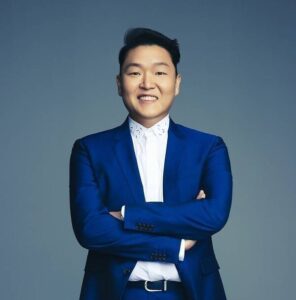 PSY poses confidently in a bright blue suit with arms crossed against a neutral background.
