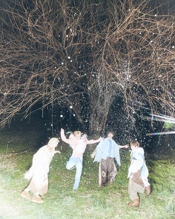 SHINee members joyfully play under a tree at night, surrounded by falling white particles that resemble snow or petals.