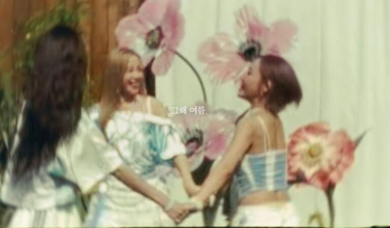 Three women hold hands in a circle outdoors, surrounded by large pink flowers and soft sunlight. Korean text on the image reads “그해 여름” (“That summer”).