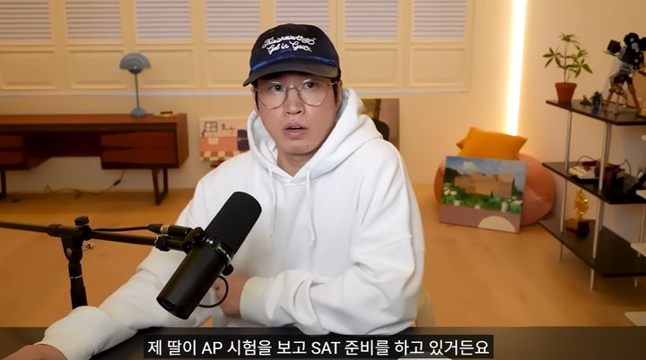Tablo speaks into a microphone during a recording session, wearing a white hoodie and cap, with Korean subtitles mentioning his daughter’s AP and SAT preparation.