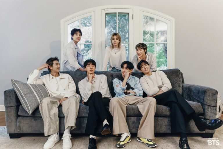 BTS members pose together on a gray sofa in a bright room with a large window behind them, dressed in light-colored casual outfits. The text “2023 FESTA BTS” appears in the corner.
