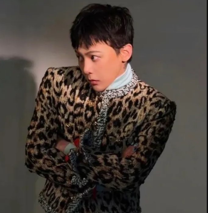 G‑Dragon poses in a leopard‑print jacket with structured shoulders, crossing his arms under dramatic lighting.