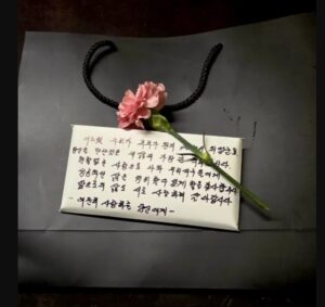A handwritten Korean letter with a pink carnation placed on top of a white envelope resting on a black surface.