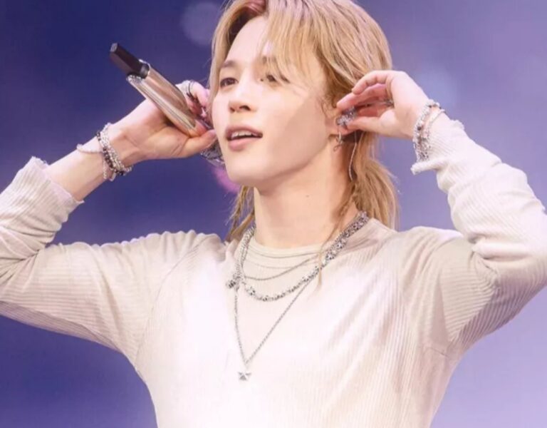 BTS’s Jimin performs on stage, holding a microphone and wearing layered silver jewelry under purple lighting.